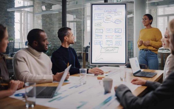 Project Manager Makes a Presentation for a Young Diverse Creative Team in Meeting Room in an Agency. Colleagues Sit Behind Conference Table and Discuss Business Development, User Interface and Design.